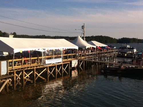 A waterfront scene with tents on a wooden dock, people gathered, and boats in the water under a clear blue sky.
