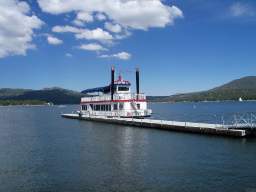 A white paddleboat docked at a pier on a calm lake, surrounded by green hills and a blue sky with fluffy clouds.