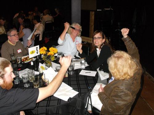 A group of people at a restaurant table, some raising their hands, with drinks and flowers on the table.