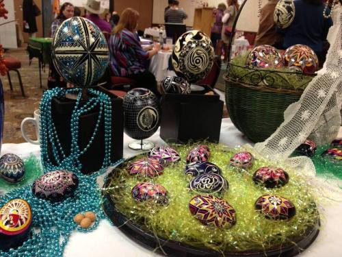 Colorful, intricately decorated eggs displayed on a table with beads and greenery, surrounded by people in a festive setting.