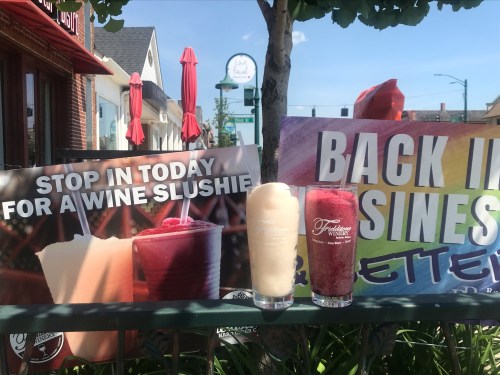 Two colorful wine slushies in clear glasses are displayed next to promotional signs outside a restaurant.