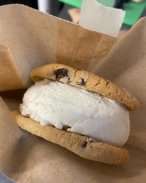 A close-up of a cookie ice cream sandwich with chocolate chip cookies and vanilla ice cream, wrapped in parchment paper.