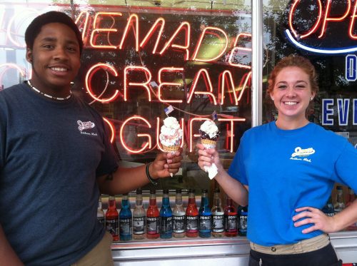 Two young people stand in front of an ice cream shop, each holding a large ice cream cone with toppings.