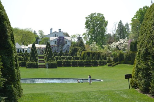 A lush garden with neatly trimmed hedges, a pond, and a child playing near the water, framed by trees and a house in the background.