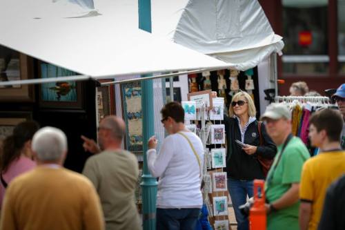 A busy outdoor market scene with people browsing art and crafts, and a woman examining items at a vendor's booth.