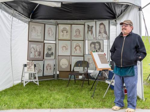 A man stands beside a tent displaying framed portraits and a sign that reads "Children, do not sit still."