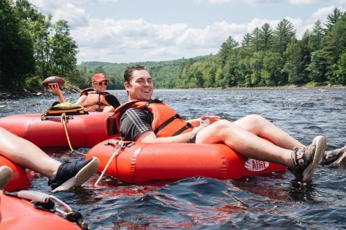 Two people in orange life jackets float on a river, enjoying a sunny day with trees and blue skies in the background.
