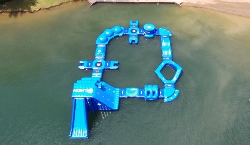 Aerial view of a large blue inflatable water park structure on a lake, featuring various slides and obstacles.