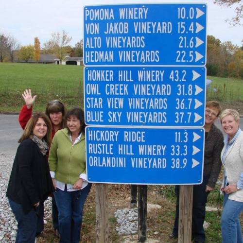 A group of six women poses by a sign listing local wineries and their distances, with a scenic background.