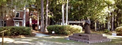 A statue of a figure stands on a brick pedestal in a wooded area, with a building and festival banner in the background.