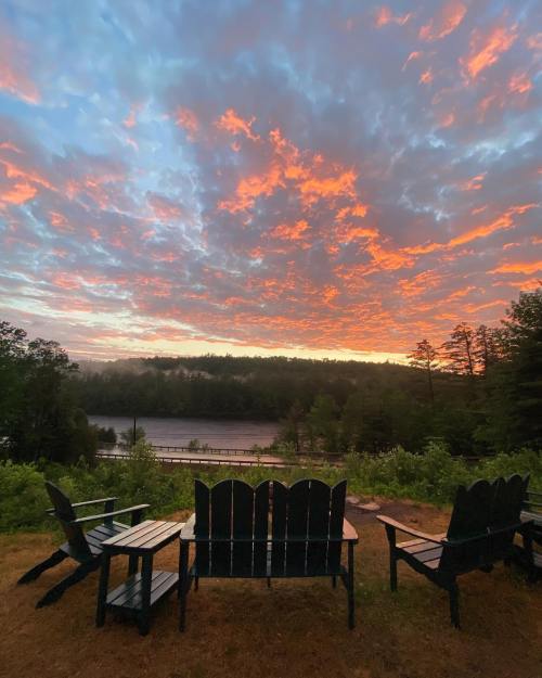 A serene lakeside view at sunset, featuring colorful clouds and wooden chairs facing the water.