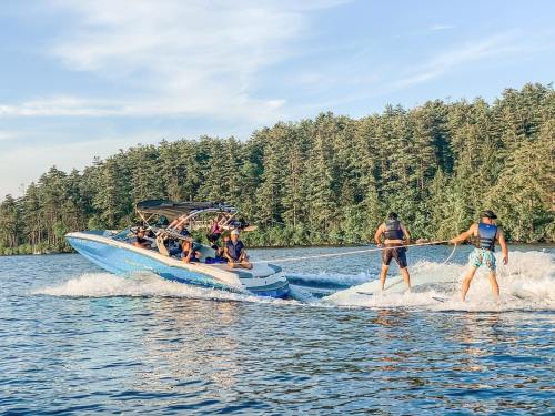 A boat with several people is towing two water skiers on a lake, surrounded by trees and a clear blue sky.