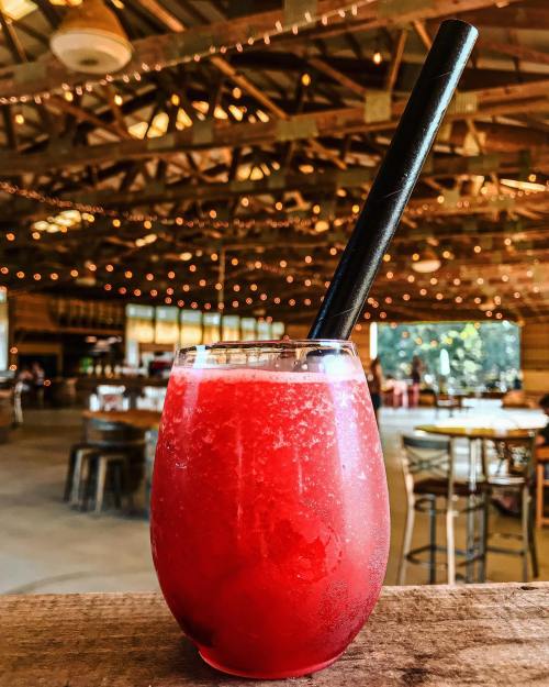 A vibrant red drink in a glass with a black straw, set against a rustic venue with string lights.
