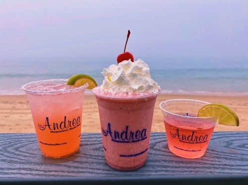 Three colorful drinks on a beachside table: a pink cocktail, a milkshake topped with whipped cream and a cherry, and another pink drink.