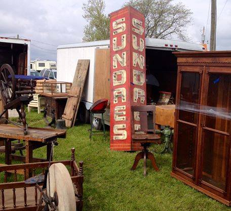 A vintage market scene featuring a tall sign reading "Sundaes" among various antique furniture and items.