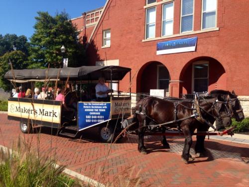 A horse-drawn carriage with passengers parked in front of a brick building, promoting Maker's Mark tours.