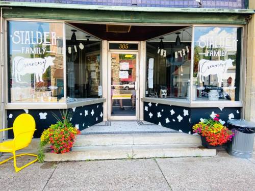 A storefront with large windows, "Stalder Family Creamery" sign, yellow chair, and colorful flower planters.