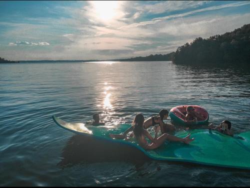 A group of children relaxes on a floating mat in a lake during sunset, with trees and a pink float nearby.