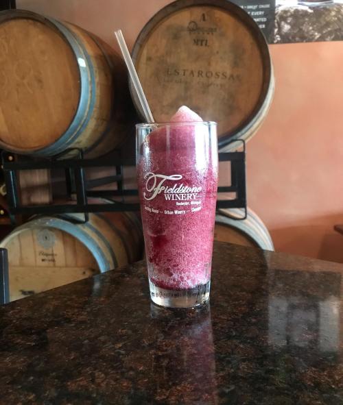 A frozen drink in a glass with a straw, set on a dark countertop near wooden wine barrels.