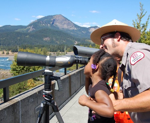 A park ranger helps a young girl look through a telescope, with mountains and a river in the background.