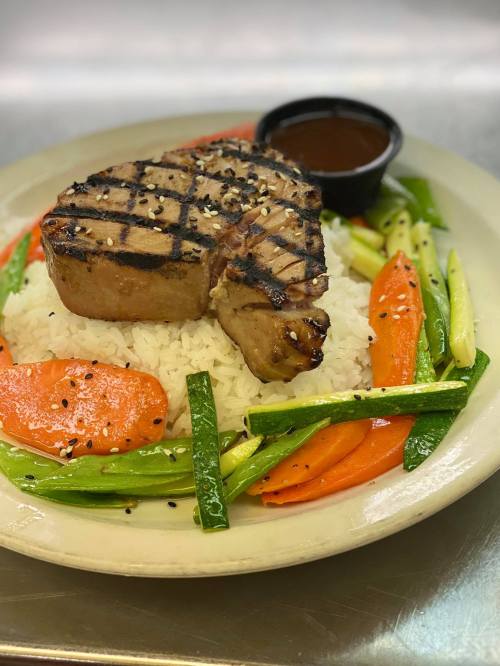 Grilled tuna steak on a bed of rice, surrounded by colorful sautéed vegetables and a side of sauce.