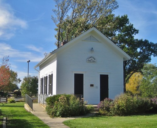 A small white town hall building surrounded by greenery and trees, with a sign reading "Camp's Town Hall."