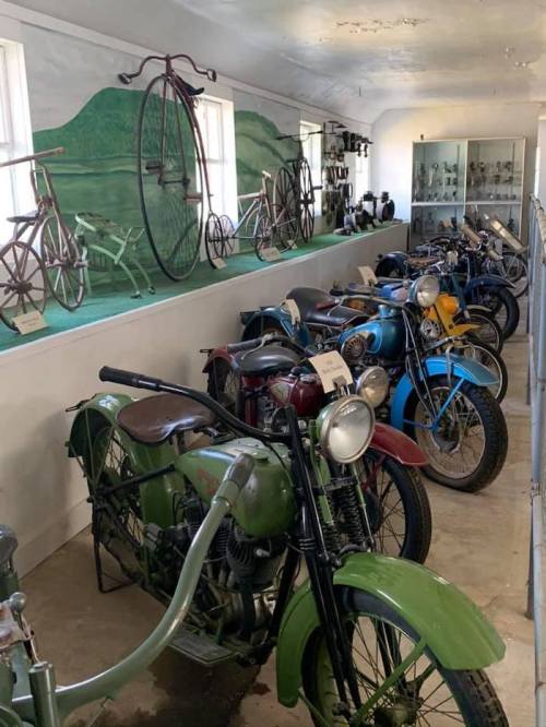 A collection of vintage motorcycles and bicycles displayed in a museum setting with green hills painted on the wall.