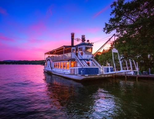 A charming riverboat docked on calm waters at sunset, with vibrant pink and purple skies in the background.