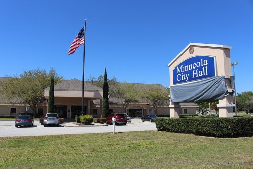 Minneola City Hall building with an American flag and a sign, surrounded by greenery and parked cars. Clear blue sky.