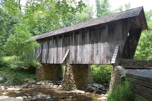 A wooden covered bridge spans a small stream, surrounded by lush greenery and trees.