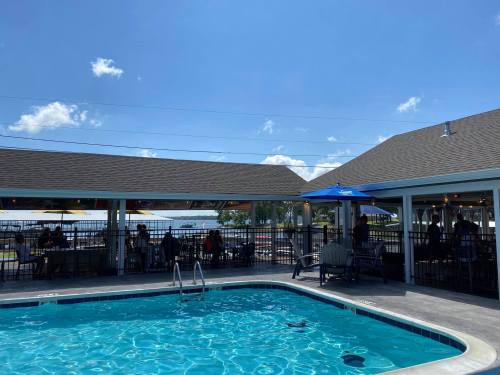 A sunny pool area with blue water, surrounded by tables and umbrellas, and a view of the lake in the background.