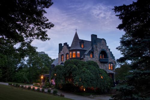 A large, stone mansion with a turret, surrounded by greenery, illuminated at dusk under a cloudy sky.