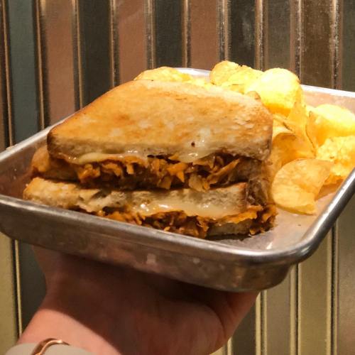 A hand holding a tray with a toasted sandwich and a side of potato chips.