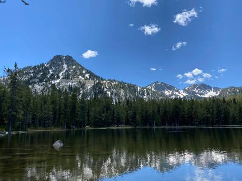 A serene lake surrounded by lush forests and snow-capped mountains under a clear blue sky with fluffy clouds.
