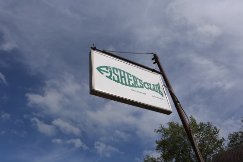 Sign for "SHEER'S CLUB" against a blue sky with clouds, hanging from a wooden post.