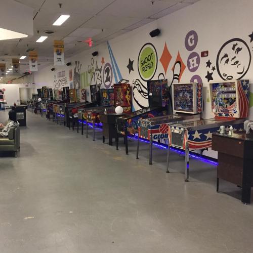 A row of colorful pinball machines lined up against a mural in a spacious arcade.