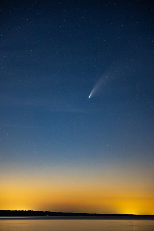 A comet streaks across a starry night sky above a calm body of water, with a gradient of colors from blue to orange.