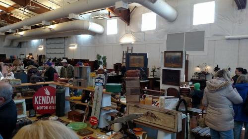 A bustling indoor market filled with vintage items, antiques, and people browsing among various stalls and displays.