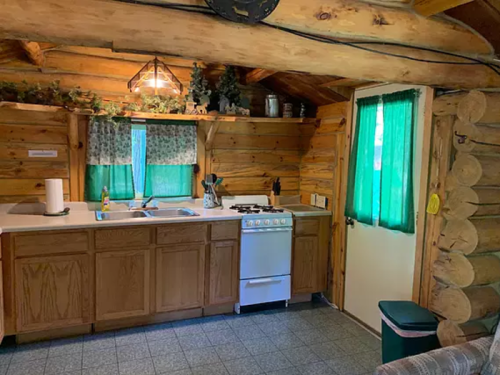Cozy log cabin kitchen with wooden cabinets, green curtains, a stove, and a sink, featuring rustic decor.