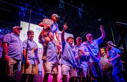 A group of eight men celebrate on stage, holding trophies and a championship belt, with one man being lifted in victory.