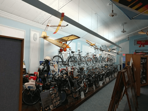 A display of vintage bicycles and model airplanes in a museum setting, with a bright blue wall and wooden flooring.