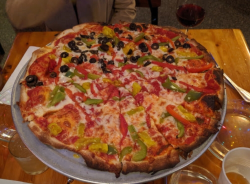 A large pizza topped with cheese, pepperoni, olives, and colorful bell peppers on a wooden table.