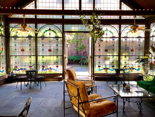 A sunlit room with stained glass windows, featuring cozy seating and a view of a garden outside.
