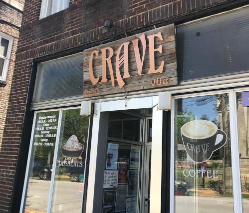 Sign for "CRAVE" coffee shop with a wooden design, featuring hours and dessert options on the storefront window.