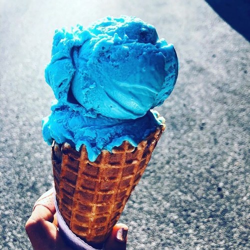A hand holding a waffle cone topped with a large scoop of bright blue ice cream against a blurred background.