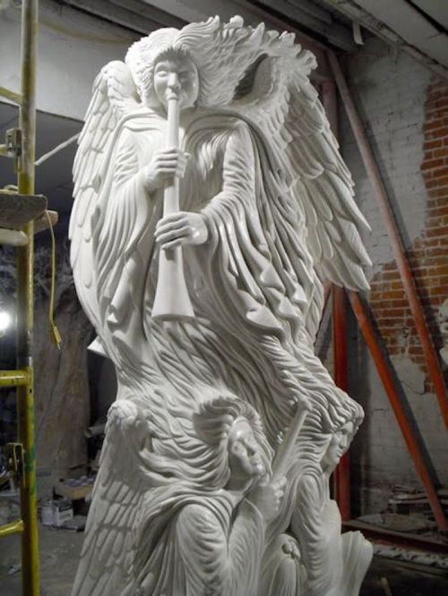A detailed white sculpture of an angel playing a trumpet, with two figures below, set in a construction environment.