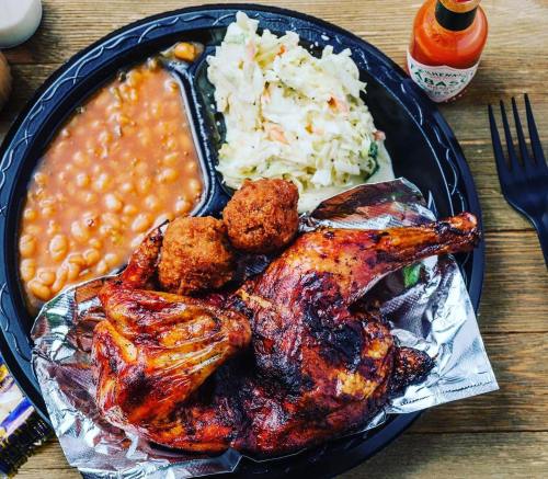 A plate featuring grilled chicken, coleslaw, baked beans, and two cornbread muffins, with hot sauce on the side.