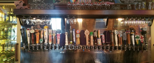 A bar with a variety of beer taps lined up, showcasing different brands and styles, with glasses hanging above.