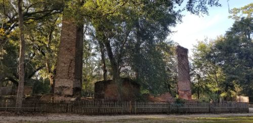 Ruins of brick structures surrounded by trees, with remnants of walls and chimneys visible in a natural setting.
