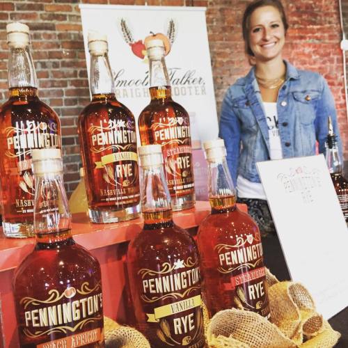 A woman stands behind a display of Pennington rye whiskey bottles at a rustic event, with a sign in the background.
